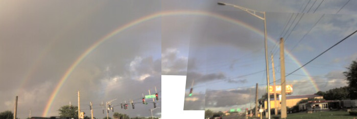 [More than four photos spliced together to show the entire rainbow. There is a white space in the middle of the image where none of the photos captured that part of the scene and overlaps of the separate photos are obvious in several places. On the left, there is a shadow of a second rainbow to the left of the clearly visible one. While the road is not visible, the traffic lights at the intersection are visible as are power lines along the right side of the road. The yellow section of the rainbow is prominently visible throughout the arc. The other colors are there, but are somewhat muted since the sun was shining brightly.]